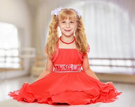 A beautiful little Caucasian girl with long, blonde ponytails on her head in a bright orange dress . Sat down on the floor.In a room with a large semi-circular window.