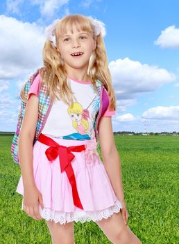 Happy little girl in a pink short skirt with a knapsack on his shoulders. Close-up.On the background of grass and blue sky with clouds.