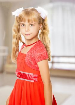 A very beautiful little girl with long, blonde ponytails on her head in a bright orange dress . close-up.In a room with a large semi-circular window.