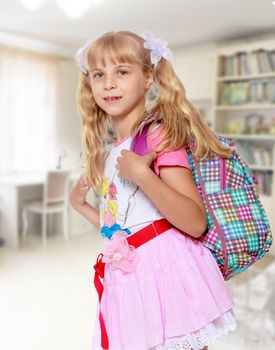 Happy little girl in a pink short skirt with a knapsack on his shoulders. Close-up.In the background children's room where there are shelves with books and toys.