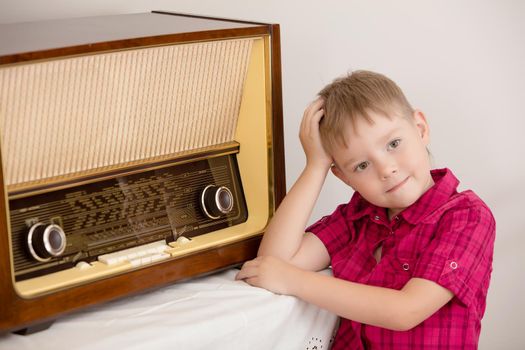 Retro style, surrounded by old things fifties of the last century. Cute little blond boy in red shirt is posing near the old radio.