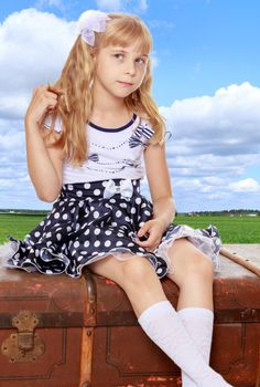Distressed little girl with long blonde ponytails on her head, sitting on the old road suitcase.On the background of bright green grass and blue sky with beautiful clouds.