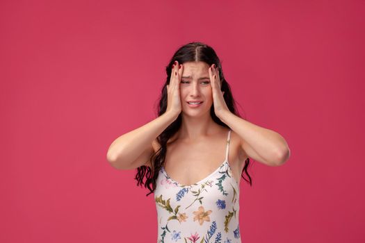 Portrait of a beautiful woman in a white dress with floral print standing on a pink wall background in studio. She act like she has a headache. People sincere emotions, lifestyle concept. Mockup copy space.