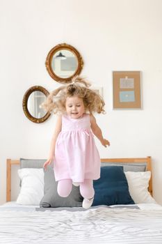 Cute little girl jumping on white bed in a pink dress