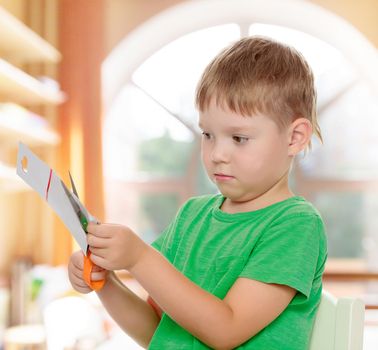 Pensive little boy cut with scissors a piece of cardboard.