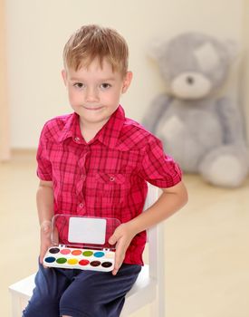 The concept of preschool development of the child ,against a child's room where in the background a Teddy bear.Cute little boy in a red shirt holds his box of watercolors.