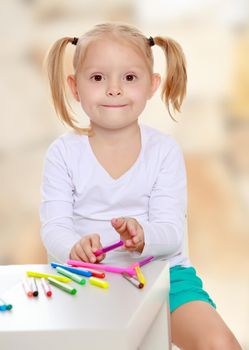 Beautiful little blonde girl with white t-shirts without a pattern, draws markers at the table on a white sheet of paper. The girl is holding a pen . close-up.