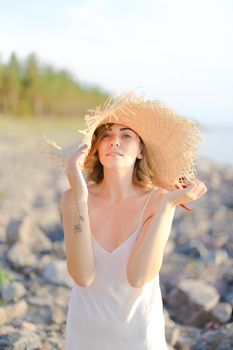 Young nice woman in hat with red ribbon standing on shingle beach. Concept of summer vacations and resort.