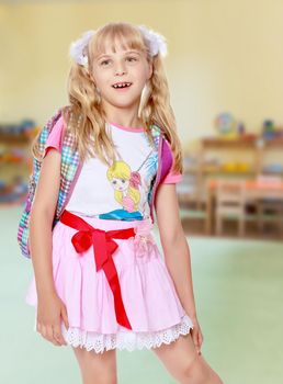 Happy little girl in a pink short skirt with a knapsack on his shoulders. Close-up.In the background children's room where there are shelves with toys.