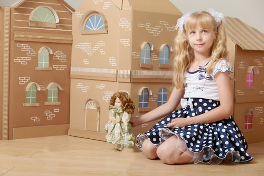 Cute little girl blonde with long ponytails on her head , playing with a doll from a cardboard small house