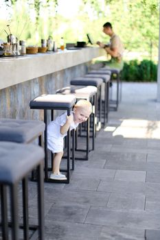 Little pretty male baby wearing white clothes and playing outside at street cafe. Concept of childhood.