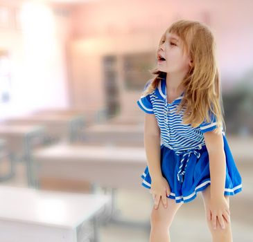 Cute little unkempt girl in a short blue dress. Girl looking to the side with his hands on his knees. In the class where there are school desks.