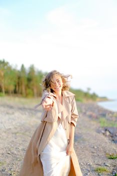 Caucasian pretty girl with outstretched hand on shingle beach wearing summer coat. Concept of seasonal fashion and summer vacations.