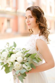 Happy caucasian fiancee keeping bouquet of flowers and wearing white dress. Concept of bridal photo session and wedding.