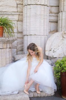 Caucasian beautiful fiancee sitting near ancient columns and and putting on shoes. Concept of bridal photo session and wedding.