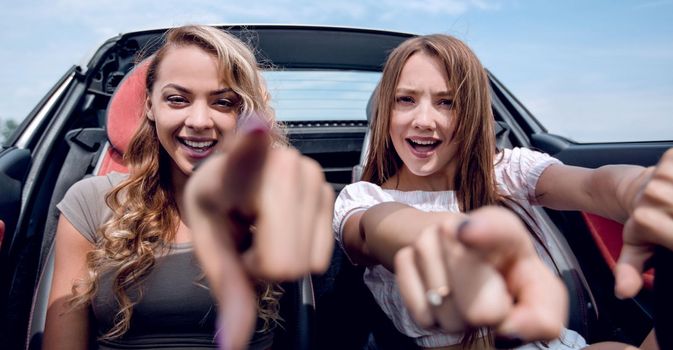 two happy girlfriends sitting in a car and pointing at you.the concept of choice