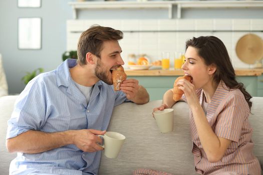 Happy young couple in pajamas in kitchen having breakfast, feeding each other a croissant