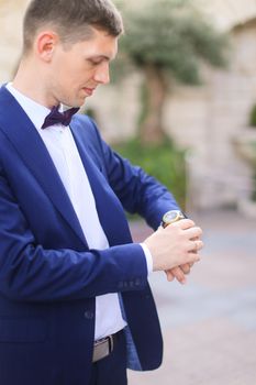 Happy european groom waiting for bridea and looking at watch, wearing dark blue suit. Concept of bridal photo session and wedding.