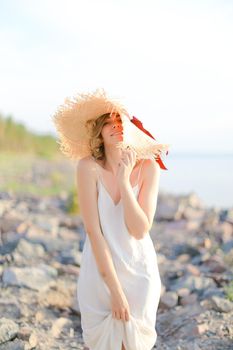 Young happy woman in hat with red ribbon standing on shingle beach. Concept of summer vacations and resort.