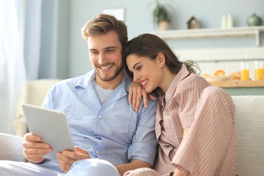 Young couple in pajamas watching media content online in a tablet sitting on a sofa in the living room