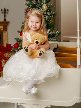 A cute little girl is sitting on a white piano with a teddy bear.
