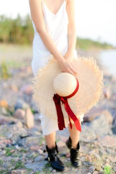 Female person standing on rocky beach with hat in hands. Concept of summer vacations and fashion.