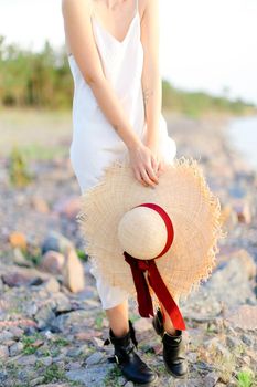 Caucasian female person standing on shingle beach with hat in hands. Concept of summer vacations and fashion.