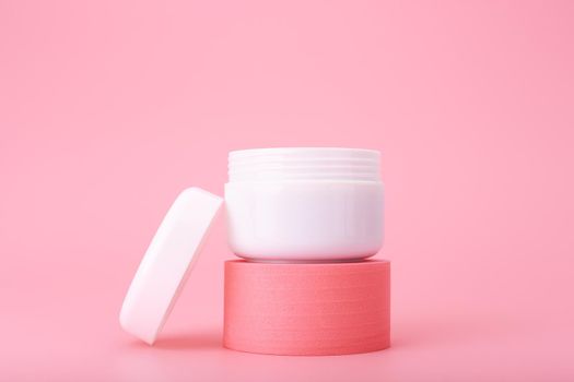 Minimal still life with white opened cosmetic jar on pedestal against pink background. Concept of repairing, moisturizing, anti aging nourishing cream, gel, mask or lotion