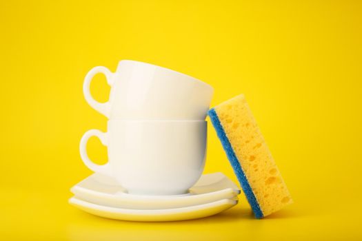 Dishwashing and housework creative minimal concept. Clean white ceramic plates, cups and yellow cleaning sponge against bright yellow background.