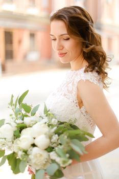 Happy nice fiancee keeping bouquet of flowers and wearing white dress. Concept of bridal photo session and wedding.