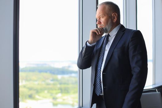 Thoughtful mature business man in full suit looking away while standing near the window