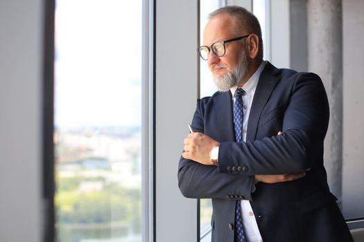 Mature businessman in a corporate suit standing in office and looking away through large windows optimistically