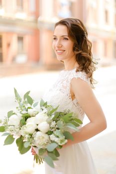 Happy european fiancee keeping bouquet of flowers and wearing white dress. Concept of bridal photo session and wedding.