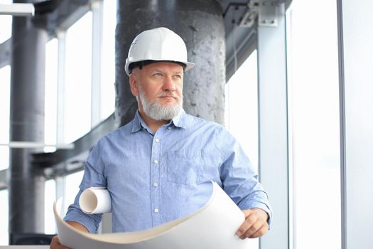 Confident mature architect in corporate suit and hardhat holding a blueprint and looking at it