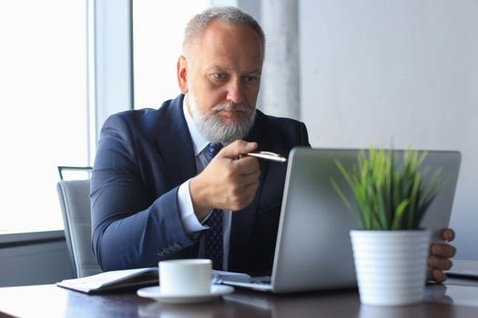 Mature businessman looking and analyzing document on laptop in his modern office at work