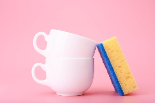 Creative, minimal dishwashing concept. Simple composition with yellow cleaning kitchen sponge next to two white ceramic cups on pink background.Close up, still life, no people