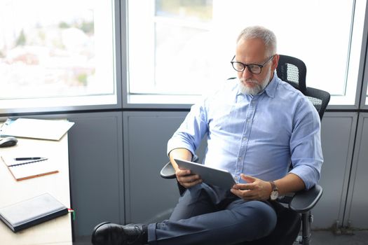 Serious mature financial advisor sitting at the table and checking financial report using digital tablet at office