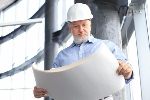 Confident mature architect in corporate suit and hardhat holding a blueprint and looking at it