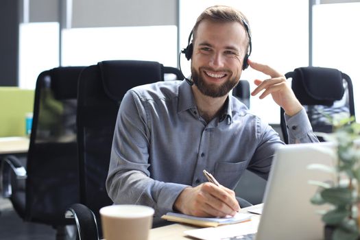 Smiling male call-center operator with headphones sitting at modern office, consulting online information in a laptop, looking up information in a file in order to be of assistance to the client