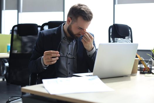 Handsome young business man with closed eyes touching face with hands while sitting on working place in office