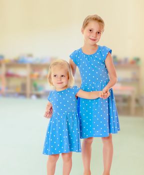 Against the background of a child's room .Two charming little girls, sisters , in identical blue dresses with polka dots , cuddling.