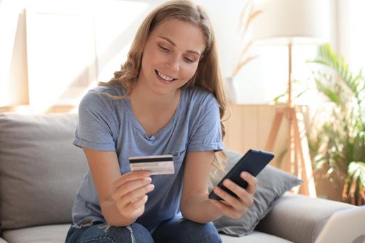 Happy natural blonde using credit card and mobile phone in living room