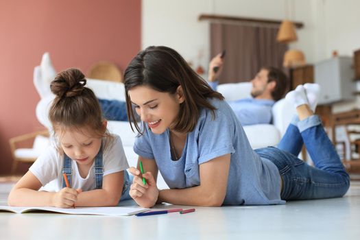 Happy mother smilling daughter lying on warm floor enjoying creative activity, drawing pencils coloring pictures in albums, father resting on couch, family spend free time together