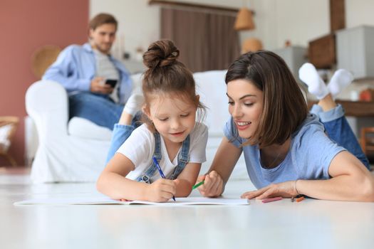 Happy mother smilling daughter lying on warm floor enjoying creative activity, drawing pencils coloring pictures in albums, father resting on couch, family spend free time together