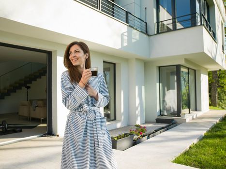 young beautiful woman in a bathrobe enjoying morning coffee in front of her luxury home villa