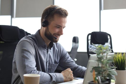 Smiling male business consultant with headphones sitting at modern office, video call looking at laptop screen. Man customer service support agent helpline talking online chat