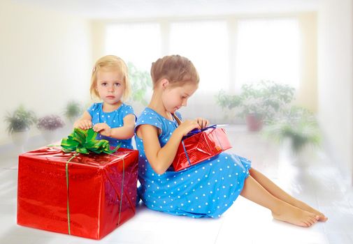 Two charming little girls , sisters, in identical blue dresses with polka dots. Girl looking at gifts Packed in beautiful red paper tied with a bow.On the background of the school hall with large Windows.