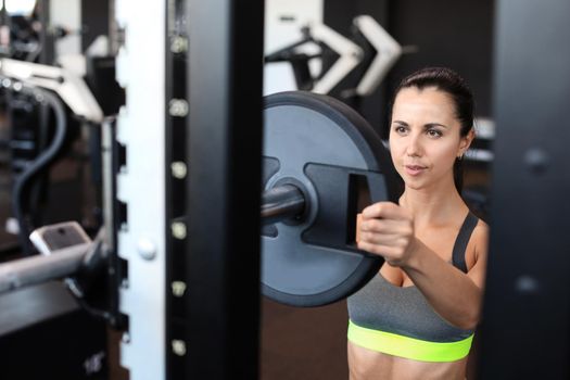 Strong young woman with beautiful athletic body adding weight on barbell at gym