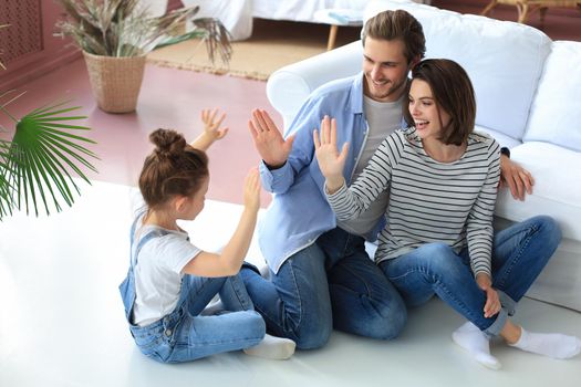 Happy young family with little child sit on warm floor relaxing together, overjoyed parents rest enjoy weekend have fun with small daughter, give high five playing