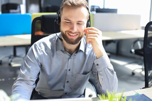 Smiling male call-center operator with headphones sitting at modern office, consulting online information in a laptop, looking up information in a file in order to be of assistance to the client